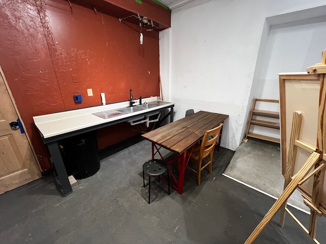 A functional room with a sink and table, possibly a kitchen or workshop area, featuring a wooden table with a black and re...