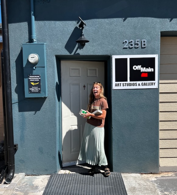 "Adult Female with Books Smiling in Front of OffMain Art Studios and Gallery"